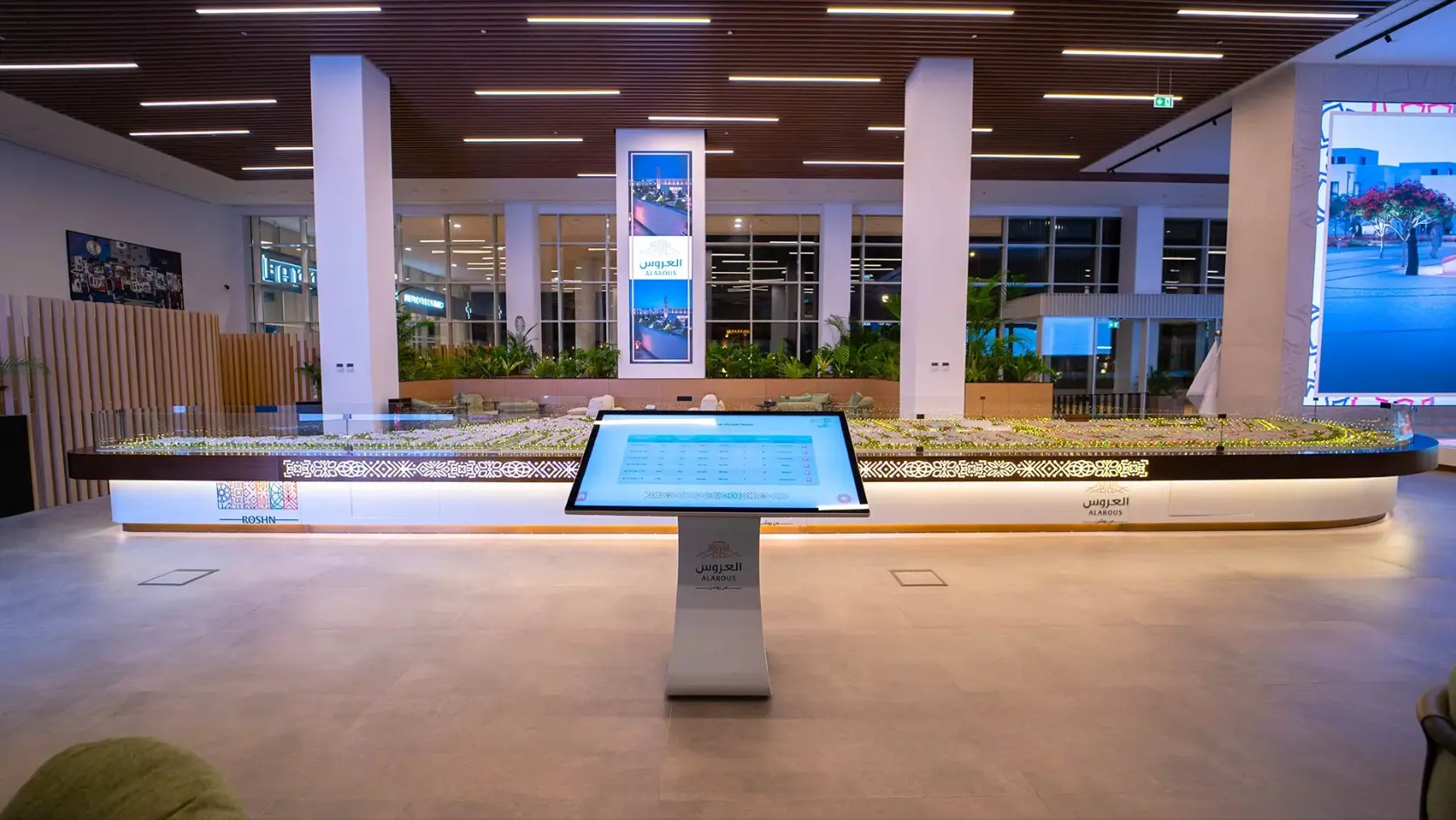 A digital display stands in front of an architectural scale model in a bright lobby.